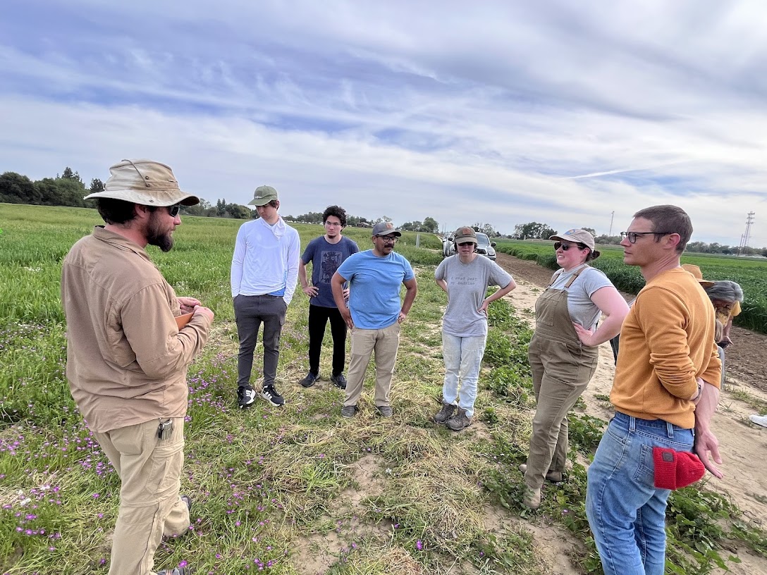 A group of seven people standing in a field, engaging in discussion, with a cloudy sky above.