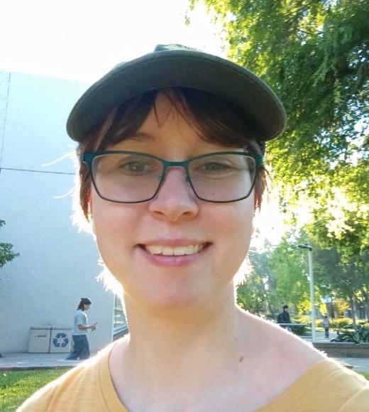 Young person wearing a cap and glasses smiles in a sunny park setting with greenery.