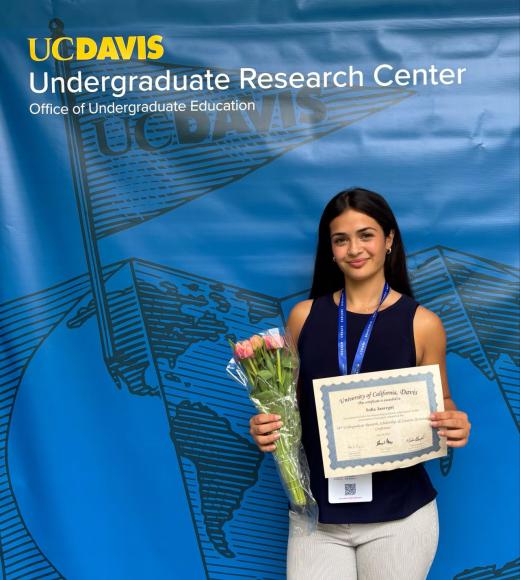 Young woman holding a certificate and flowers in front of a UC Davis banner.