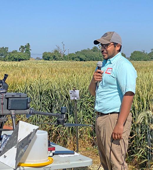 A man in a cap stands by a table with equipment, speaking in a field of crops.