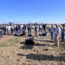 People gathered around equipment in a sunny agricultural field under blue sky